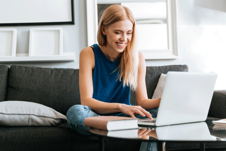 imagem central de uma mulher branca, de cabelo loiro, mexendo em seu notebook. A mulher usa uma regata azul-escuro e uma calça jeans com a barra dobrado, e está de pés descalços, sentada em um sofá na cor grafite. À sua frente, há uma pequena mesa de vidro com o notebook, um livro com capa na cor rosé e uma folha. Em cima do sofá há uma almofada cinza e ao fundo é possível avistar uma parede branca com quadros nas cores branco e preto.