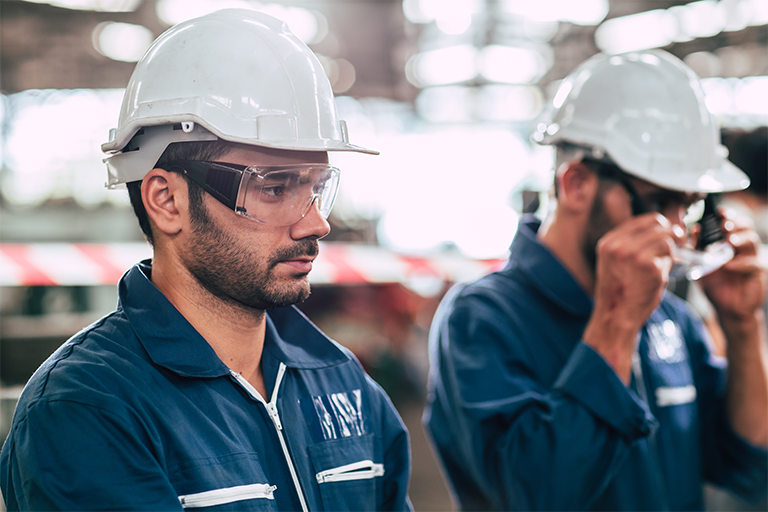 A imagem exibe, em uma visão lateral, dois homens trabalhadores da indústria, posicionados um ao lado do outro. O homem em primeiro plano é branco, possui barba preta curta e usa um uniforme na cor azul-escuro, capacete branco e óculos de proteção individual. O homem à direita da imagem, embaçado, usa os mesmos equipamentos e está colocando os óculos na face.