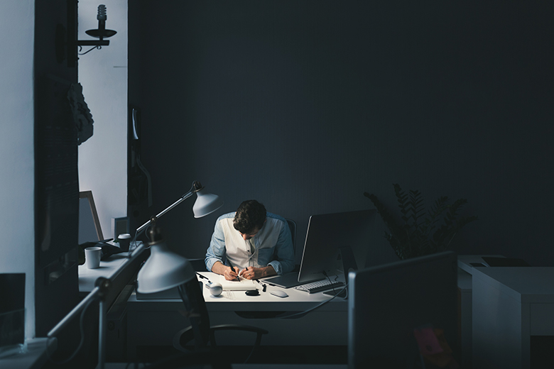 Imagem de um homem de cabelo escuro e camisa jeans, sentado em frente a uma mesa de trabalho. Ele está com a cabeça voltada para a mesa, onde escreve em um papel. O ambiente é escuro e mais à frente da imagem é possível visualizar outra mesa de trabalho com uma tela de computador.