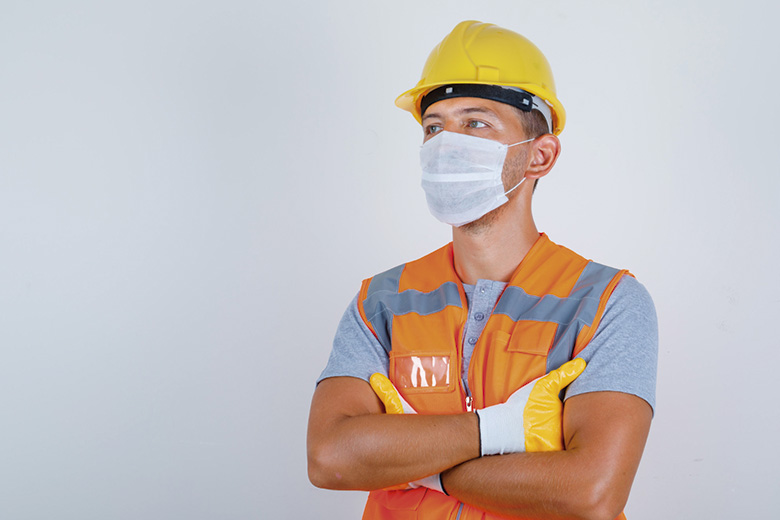 A imagem mostra um homem branco, de olhos claros, posicionado à direita. Ele está usando uniforme de construtor: camiseta cinza com colete laranja com cinza por cima, máscara de proteção branca, capacete amarelo e luvas amarelo com branco. Seus braços estão cruzados e seu olhar voltado para a esquerda. O fundo da imagem é cinza.