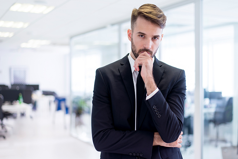 A imagem mostra um homem branco, de cabelo e barba castanhos, usando um terno preto. Ele está posicionado à direita da imagem com a mão esquerda tocando seu queixo em posição de dúvida. Ao fundo, é possível visualizar mesas de trabalho em plano embaçado.