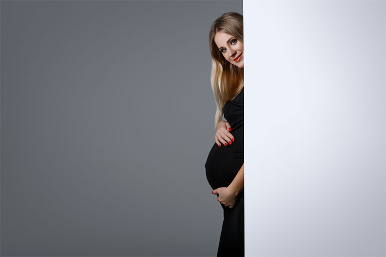 Na imagem, uma mulher jovem, de pele clara, cabelos loiros longos e olhos azuis está usando um vestido preto. Ela está grávida e, de forma sutil, posiciona suas mãos sobre a barriga.