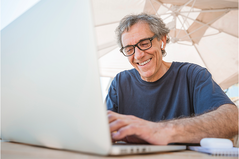 A imagem mostra um homem branco, de cabelos grisalhos, teclando em um notebook. Ele veste camiseta azul marinho, óculos de grau com armação preta e está sorrindo enquanto olha para a tela do notebook, que é cinza. Atrás dele, em segundo plano, há um guarda-sol em tons de marrom e bege.