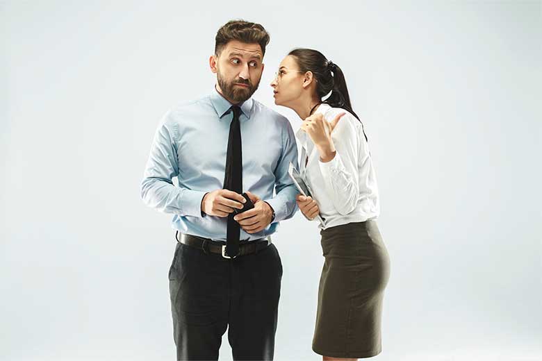 Homem branco de terno com aspecto de surpresa ouvindo uma moça branca, com camisa e saia social, que lhe conta algo, em tom de suspense, próximo ao seu ouvido.