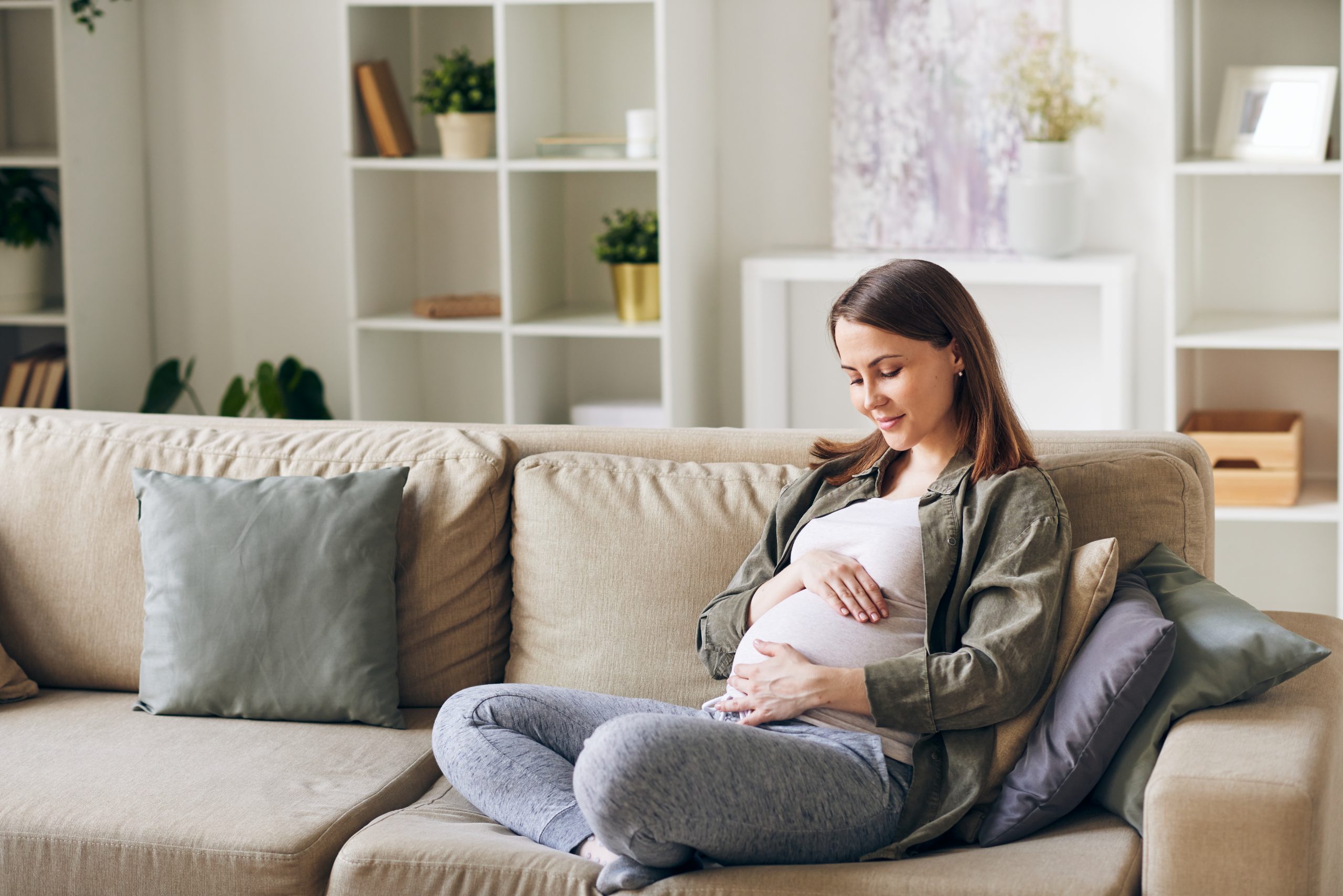 A imagem mostra uma mulher caucasiana, grávida, sentada em um sofá bege. Ela veste calça jeans, blusa branca e camisa verde, e com as duas mãos toca sua barriga, enquanto a observa. Em suas costas, estão três almofadas em tons pasteis e, à sua direita, há mais uma almofada, cinza. Ao fundo, é possível visualizar algumas prateleiras brancas com dois vasos de flor.