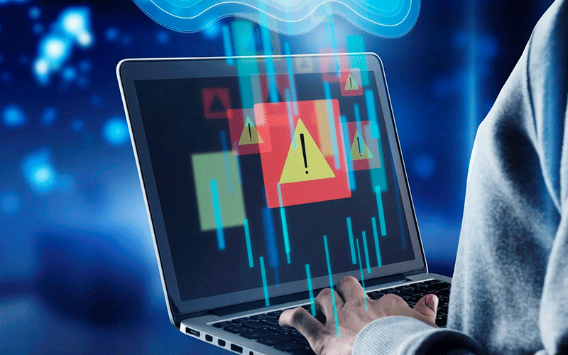 A imagem mostra um cenário cibernético, com um notebook. Em sua tela, há sinais de alerta a respeito do armazenamento de dados tóxicos na empresa. Sob o notebook há uma imagem holográfica da nuvem de armazenamento. Mexendo no computador há um indivíduo usando roupas cinzas. Apenas parte do seu tronco e mão podem ser vistos.